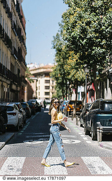 Young stylish woman with drink in hand crossing street