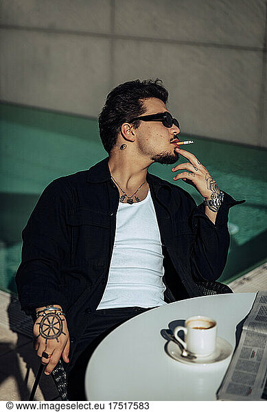 Young tattooed guy smoking a cigar on a terrace while having a coffee