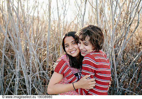 Young teen sisters hug cheek to cheek and smile in a wooded area