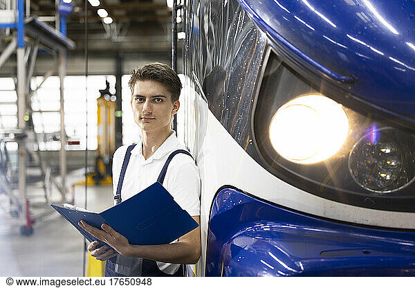 Young trainee with file folder standing by monorail at industry