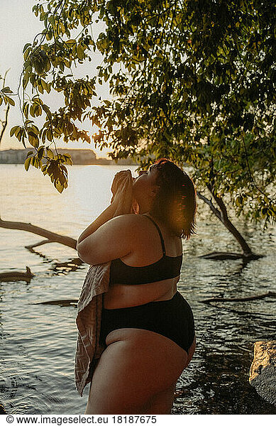 Young voluptuous woman with towel standing in water against tree during sunset