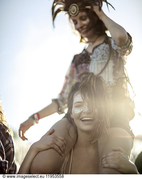 Young woman at a summer music festival carrying a friend on her shoulders  face painted  smiling at camera.