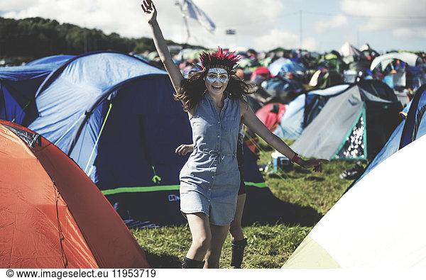 Young woman at a summer music festival face painted  wearing feather headdress  standing near the campsite surrounded by tents  arm raised  smiling.