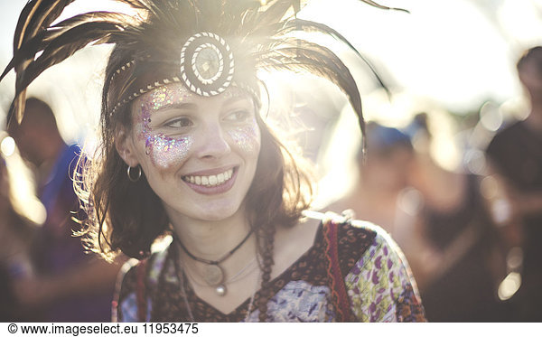 Young woman at a summer music festival wearing feather headdress and face painted  smiling.