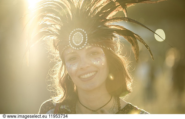 Young woman at a summer music festival wearing feather headdress and face painted  smiling at camera.