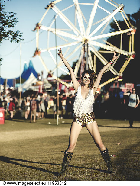 Young woman at a summer music festival wearing golden sequinned hot pants  face painted  smiling at camera  arms raised.