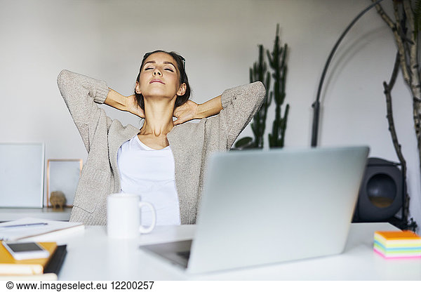 Young woman at home with laptop on desk leaning back