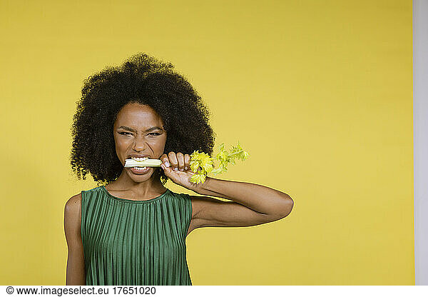 Young woman biting celery against yellow background