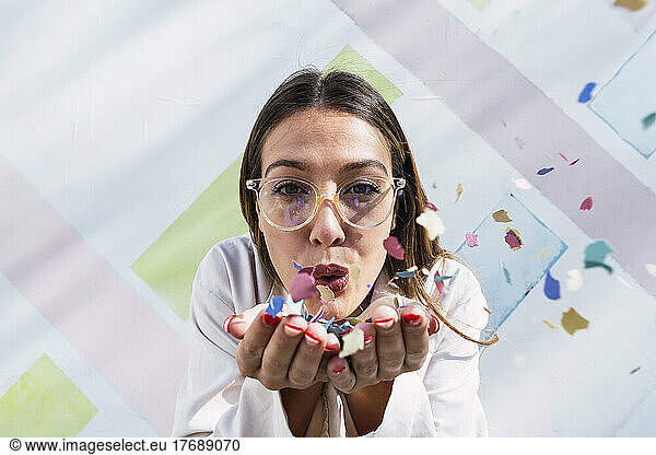 Young woman blowing multi colored confetti in front of wall