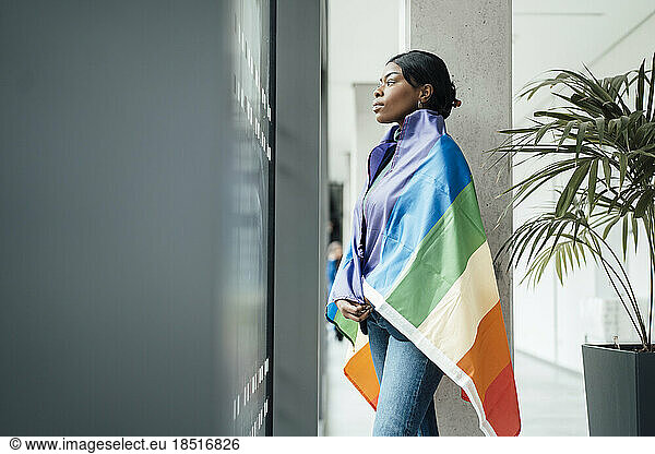 Young woman covered in rainbow flag looking out of window at office