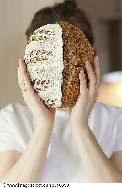 Young woman covering face with loaf of bread at home