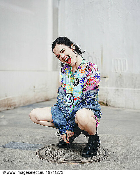 Young woman crouching on manhole and laughing in front of white wall
