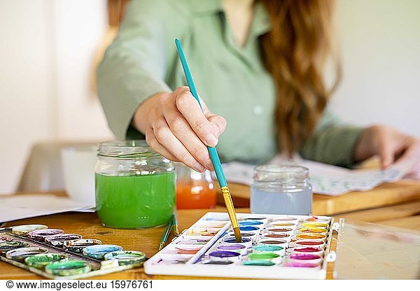Young woman dipping paintbrush in watercolor on table at home