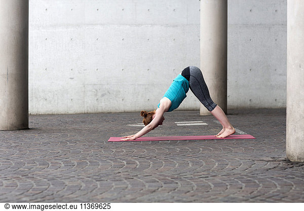 Young woman doing downward facing dog pose on exercise mat in urban city