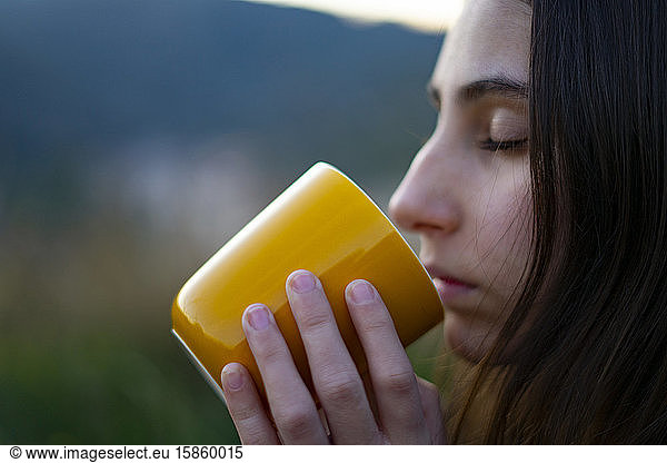 Young woman drinking and smelling coffee in a yellow cup in the woods.