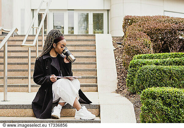 Young woman drinking coffee from reusable mug while sitting on steps