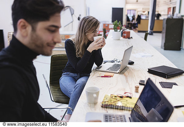Young woman drinking coffee while sitting with colleague at desk in office