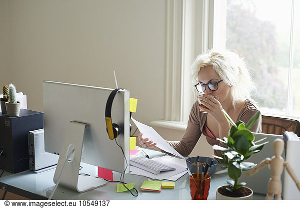 Young woman drinking tea and reading paperwork at desk in home office