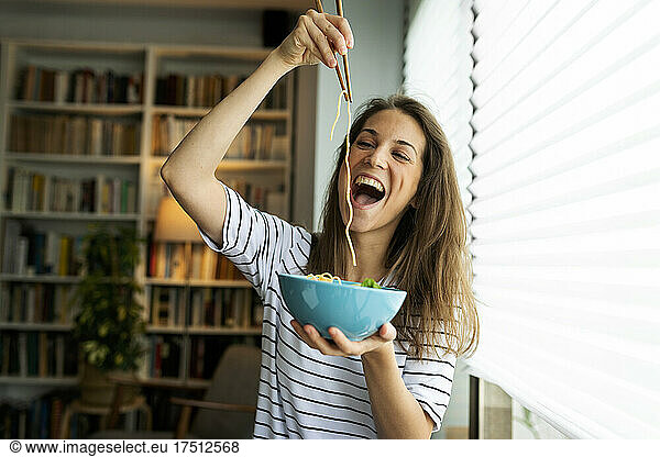 Young woman eating spaghetti while sitting by window at home