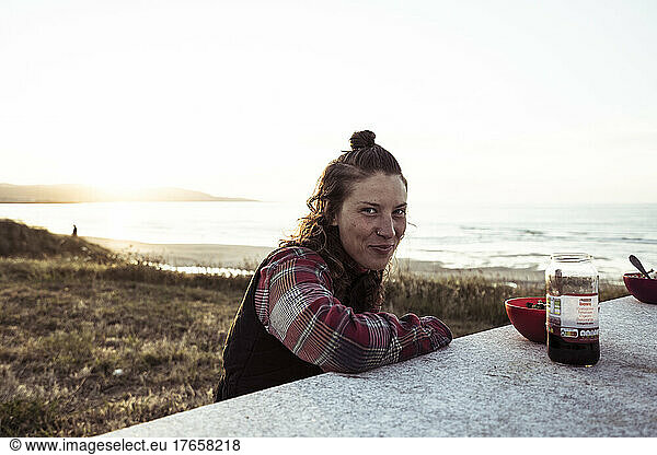Young woman eats food near ocean and setting sun