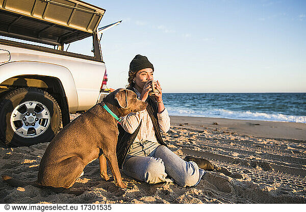 Young woman enjoying drink in mug while beach car camping with dog