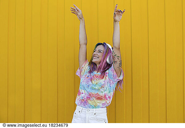 Young woman enjoying music listening through wireless headphones and dancing in front of yellow wall