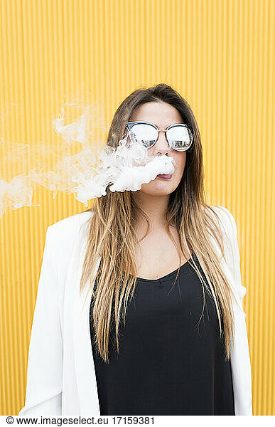 Young woman exhaling smoke against yellow wall