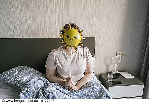 Young woman hiding face with smiley balloon on bed at home