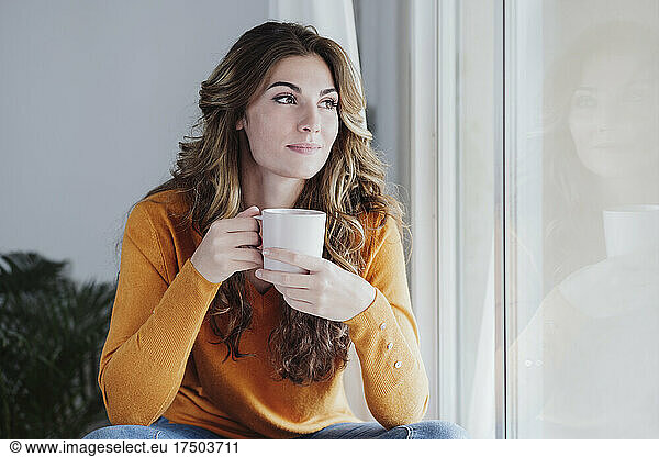 Young woman holding coffee cup by window at home