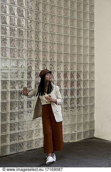 Young woman holding coffee cup looking away while standing against glass wall