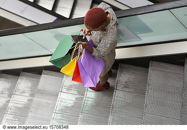 Young woman holding colorful shopping bags and checking her cell phone while descending the stairs