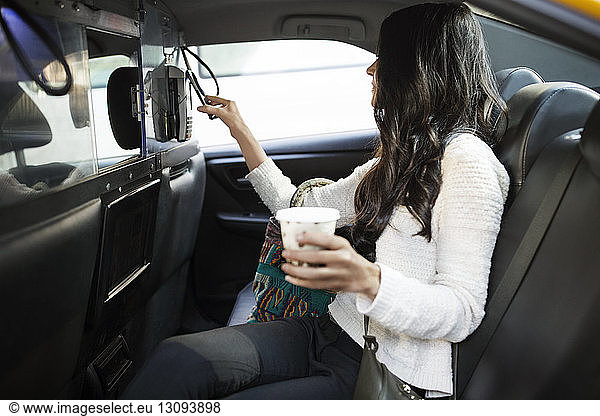 Young woman holding disposable glass while making contactless payment through smart phone in taxi