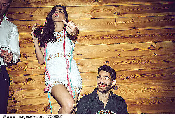 Young woman holding drink showing middle finger while enjoying with male friends in party
