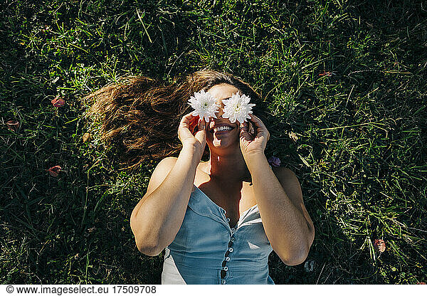 Young woman holding flowers over face while relaxing on grassy land in park