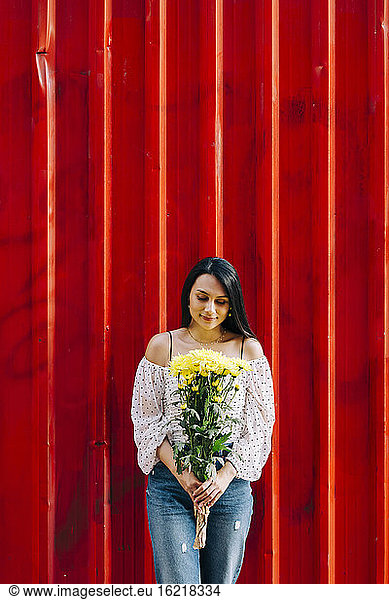 Young woman holding flowers while standing against red wall