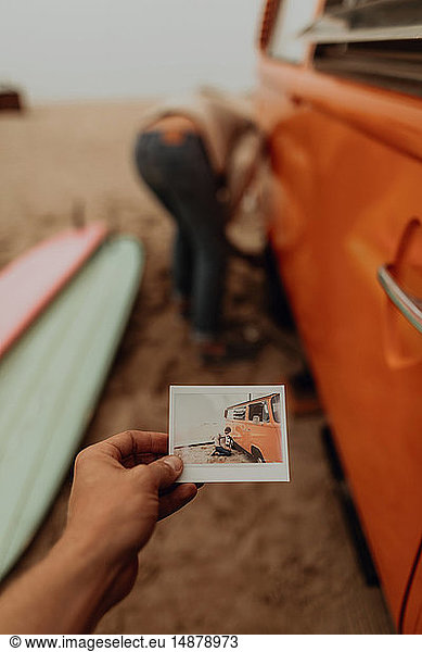 Young woman holding instant photo of boyfriend removing flat tyre on recreational vehicle at beach  Jalama  California  USA