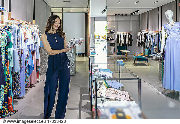 Young woman holding purse while standing near clothes rack in boutique