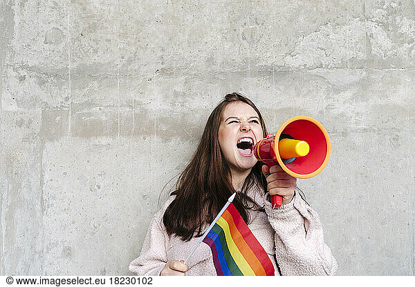Young woman holding rainbow flag shouting through megaphone leaning on wall