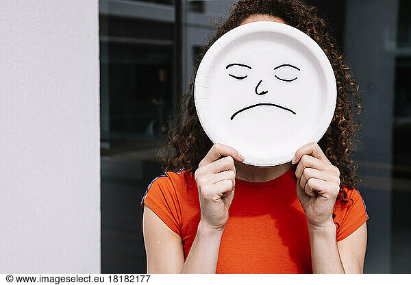 Young woman holding sad emoticon plate over face