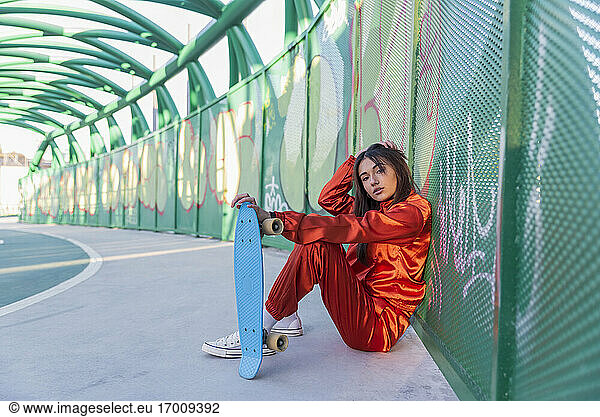 Young woman holding skateboard while sitting with hand in hair on bridge