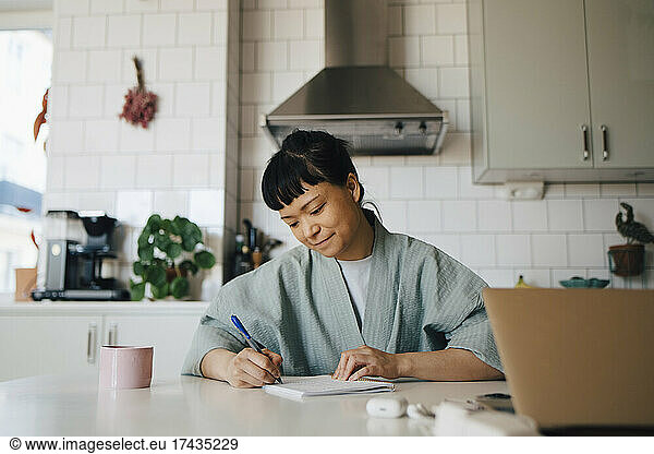 Young woman in bathrobe writing in book at domestic kitchen