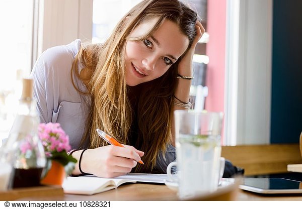 Young woman in cafe writing  hand on head resting on elbow looking at camera smiling