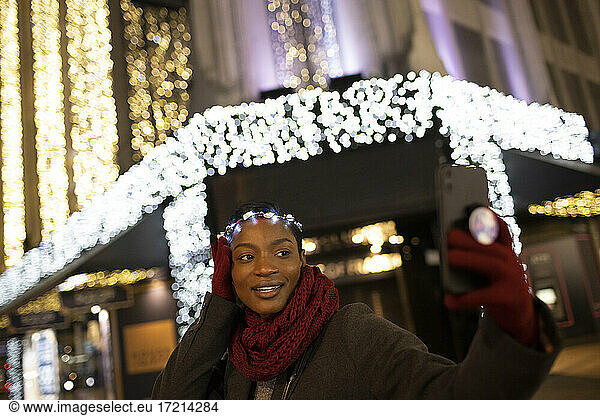 Young woman in Christmas light headband taking selfie in city at night