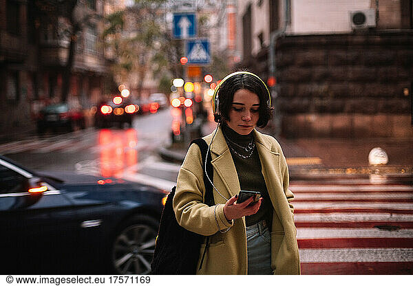 Young woman in headphones using smart phone standing on street in city