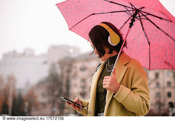 Young woman in headphones with an umbrella using smart phone in city
