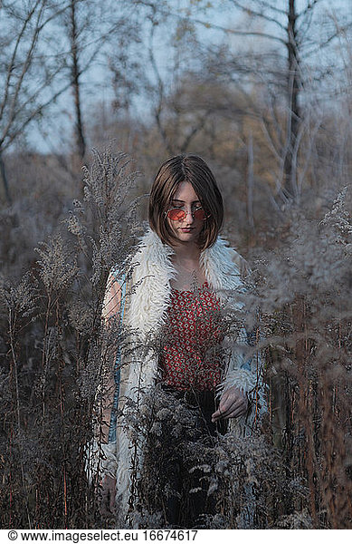 Young woman in hippie coat and red sunglasses among weeds and gr