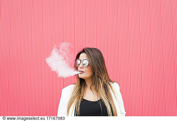 Young woman in sunglasses exhaling smoke against pink wall