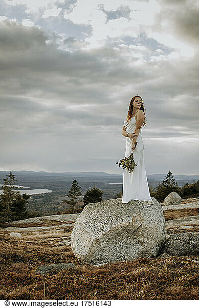 Young woman in wedding dress stands on large boulder on mountain