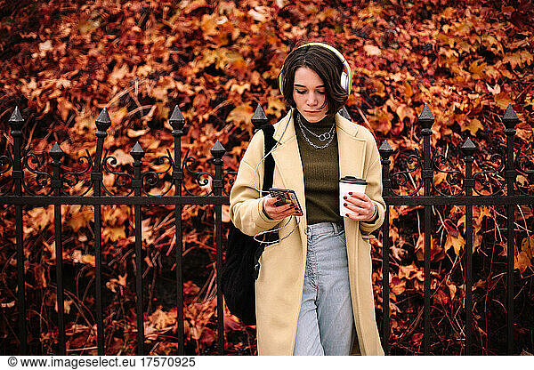 Young woman listening music in headphones standing in city in autumn