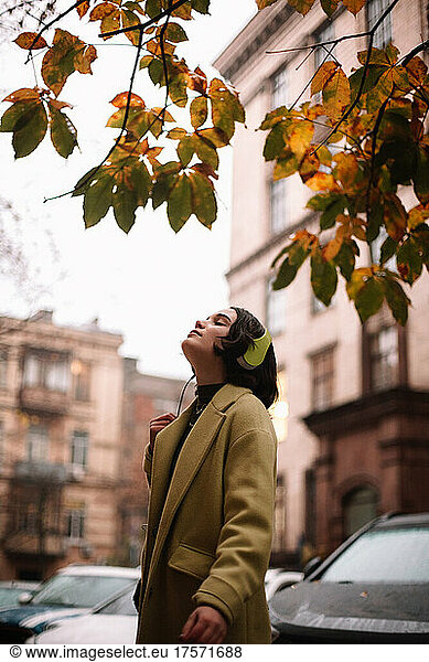 Young woman listening music in headphones standing on street in autumn